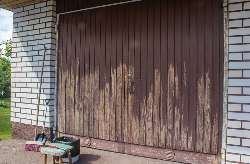 Garagedeuren van hout met gedeeltelijk afgebladderde verf en voorbereide schoonmaakbenodigdheden ernaast.